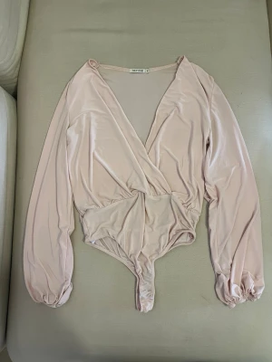 Ljusrosa omlottbody med långa ärmar - Säljer en ljusrosa body från NLY ONE med omlott framtill och långa puffiga ärmar. Bodyn har resår vid ärmsluten och är gjord i ett mjukt, glansigt material som ger en elegant look. Perfekt att styla till jeans eller kjol.