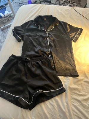 Svart satinpyjamas med vita detaljer - Snygg svart pyjamas i glansigt satinmaterial med vita kontrastkanter. Setet består av en kortärmad skjorta med knappar och matchande shorts med resår i midjan. Perfekt för mysiga kvällar hemma.