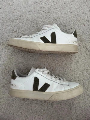 Vita Veja sneakers med gröna detaljer - Säljer ett par klassiska vita Veja sneakers med gröna mockadetaljer på hälen och det ikoniska V:et på sidan. Skorna har lågt skaft, vita snören och en beige sula. Tillverkade i läder och mocka för en clean och stilren look.