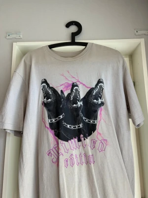 Beige t-shirt med cerberus-tryck - Säljer en beige t-shirt med stort tryck av tre svarta hundhuvuden (cerberus) och texten 'Limited edition' i lila. T-shirten har rund hals och korta ärmar. Perfekt för dig som gillar unika och edgy motiv.