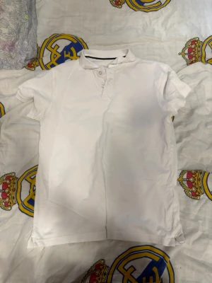 Vit kortärmad pikétopp - Klassisk vit pikétopp med korta ärmar och knappar vid halsen. Enkel och stilren design som passar till det mesta. Tillverkad i mjukt bomullsmaterial för en bekväm känsla.