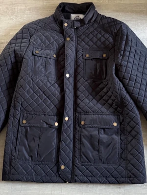 Saint Vincent Svart quiltad jacka med fickor - Snygg svart quiltad jacka Saint Vincent med fyra framfickor och tryckknappar. Jackan har hög krage och lång ärm, perfekt för kyligare dagar. Klassisk design med rutmönster och detaljer i guld på knapparna. Tillverkad i polyester för en lätt och bekväm känsla.