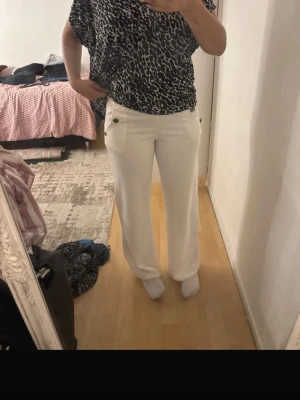 Vita kostymbyxor med knappar - Säljer ett par vita kostymbyxor med raka ben och hög midja. Byxorna har dekorativa svarta knappar vid fickorna framtill och en stilren look. Perfekta för dig som vill ha en clean och modern stil. Materialet är dock ej kostym liknande utan skönare material.