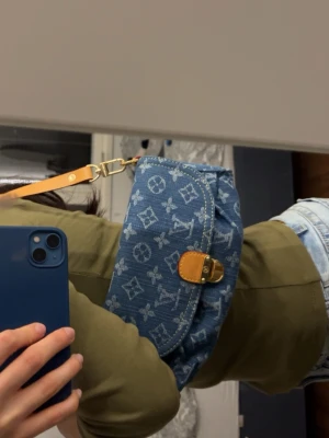 Blå väska - Säljer en snygg blå axelväska i denim med vitt monogrammönster och guldfärgat spänne. Väskan har en justerbar rem i ljust läder och en praktisk ficka framtill. Perfekt för dig som vill ha en trendig och unik accessoar. Skriv för mer info💗