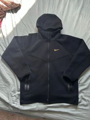Svart Nike hoodie med dragkedja - Säljer en svart hoodie från Nike med hel dragkedja framtill och gul Nike-logga på bröstet. Hoodien har två fickor med dragkedja och en stor huva. Materialet är tjockt och mjukt, perfekt för chill dagar. Passar dig som gillar sportig och clean stil. Står st M fast passar som S