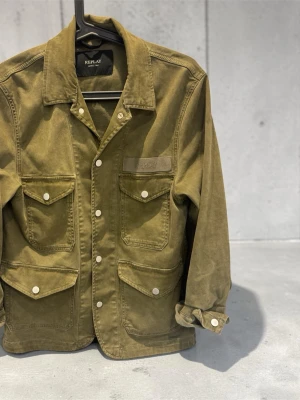 Olivgrön overshirt från Replay - Stilren olivgrön overshirt från Replay med fyra stora fickor framtill och vita tryckknappar. Klassisk krage och raka ärmar med knapp vid ärmslut. Tillverkad i slitstark bomull, perfekt för lager på lager och en avslappnad look. Köpt i butik för 2899kr 