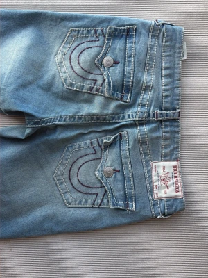 Ljusblå jeans från True Religion - Snygga ljusblå jeans från True Religion med klassiska kontrastsömmar och ikoniska bakfickor med lock och knapp. Jeansen har en bootcut passform och är tillverkade i mjuk denim med lätt tvättad look. Perfekta för en avslappnad stil.