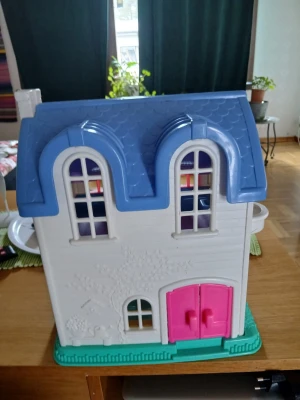 Dockhus Fisher-Price blått tak - Säljer ett klassiskt dockhus från Fisher-Price med blått tak, vita väggar och rosa detaljer runt fönster och dörrar. Huset har flera fönster, en rosa dörr och står på en grön bas. Perfekt för lek och fantasi!