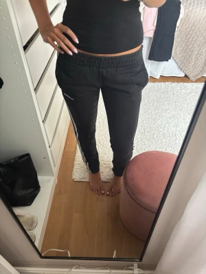 Svarta Adidas mjukisbyxor med vita ränder - Säljer ett par svarta mjukisbyxor från Adidas med klassiska vita ränder längs sidorna och Adidas-logga på vänster ben. Byxorna har resår i midjan och muddar vid bensluten. Perfekta för chill dagar eller träning.