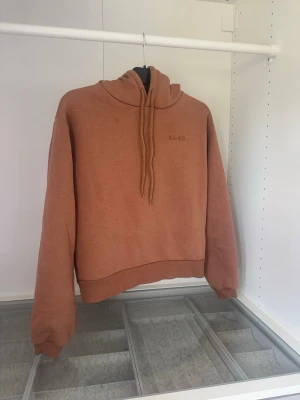 Rostbrun hoodie från NA-KD - Säljer en snygg rostbrun hoodie från NA-KD med huva och dragsnören. Hoodien har en relaxed fit, ribbade muddar vid ärmslut och nederkant samt diskret logga på bröstet. Perfekt för en chill och trendig look.