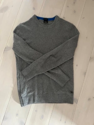 Grå stickad tröja från Hugo Boss - Mysig grå stickad tröja från Hugo Boss med rund halsringning och ribbade muddar vid ärmslut och nederkant. Tröjan har en enkel och stilren design, perfekt för lager på lager och passar till det mesta. Storlek M men känns mer som S.