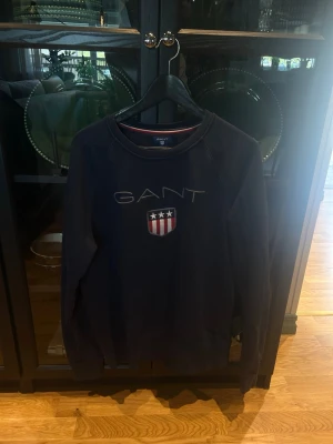 Mörkblå sweatshirt från GANT XL - Mörkblå sweatshirt från GANT med broderad logga och sköld med stjärnor och ränder på bröstet. Tröjan har rund halsringning och ribbade muddar vid ärmslut och nederkant. Klassisk och stilren design med sportig känsla. Går att pruta