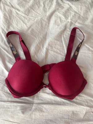 Vinröd push-up bh med strassdetaljer - Snygg vinröd push-up bh med vadderade kupor och breda axelband dekorerade med glittrande strass och guldfärgade loggor. Bh:n har slät yta och justerbara axelband för bästa passform. Endast använd 1 gång så är så gott som ny 💕Jag har vanligtvis 70B ifall ni är osäkra på måttet vilket passar jättebra! 