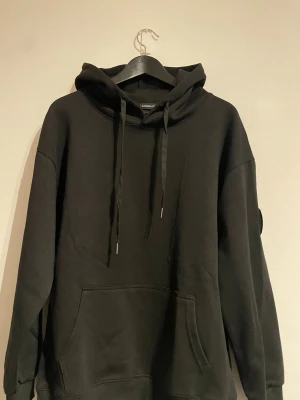 Svart hoodie från Canada Goose - Säljer en svart hoodie från Canada Goose med klassisk huva och dragsnören. Hoodien har en stor magficka och en snygg patch på ärmen. Tillverkad i mjukt material som känns skönt mot huden. Perfekt för dig som gillar stilrena och bekväma plagg.