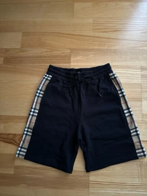 Svarta Burberry shorts med rutiga detaljer - Snygga svarta shorts från Burberry med klassiska rutiga paneler längs sidorna. Shortsen har dragsko i midjan och är tillverkade i mjukt bomullsmaterial. Perfekta för en avslappnad och trendig look.