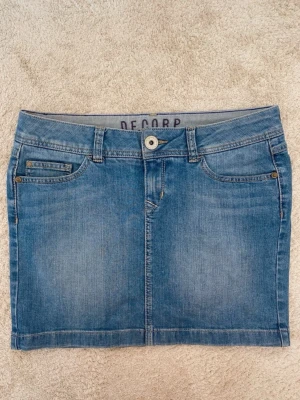 Blå jeanskjol från Decorp - Snygg blå jeanskjol från Decorp med klassisk femficksdesign, bälteshällor och knappstängning fram. Kjolen har en rak passform och är tillverkad i slitstarkt denimtyg. Perfekt till vardags för en avslappnad look.
