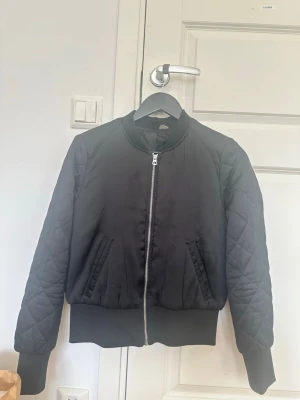 Svart bomberjacka med quiltade ärmar - Stilren svart bomberjacka med quiltade ärmar och breda muddar. Jackan har två snedställda fickor framtill och en silvrig dragkedja. Materialet är glansigt och ger en modern look. Perfekt för dig som vill ha en klassisk men trendig jacka.