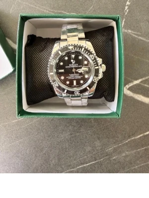 Rolx Submariner silver herrklocka - Stilren Rolx Sub herrklocka i silver med svart urtavla och tydliga vita markeringar. Klockan har en klassisk rund form och länkarmband i metall. Perfekt accessoar för dig som gillar tidlös design och exklusiv känsla.