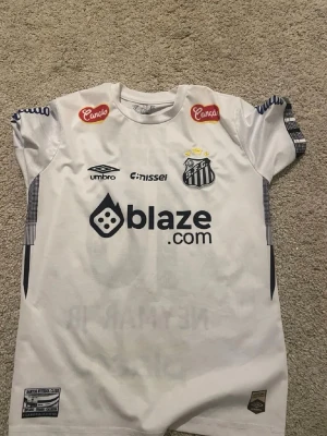 Vit Santos fotbollströja Neymar Jr - Säljer en vit Santos fotbollströja från Umbro med Neymar Jr och nummer 10 på ryggen. Tröjan har flera sponsorloggor, rund hals och korta ärmar. Perfekt för dig som älskar brasiliansk fotboll och vill ha en ikonisk supportertröja.
