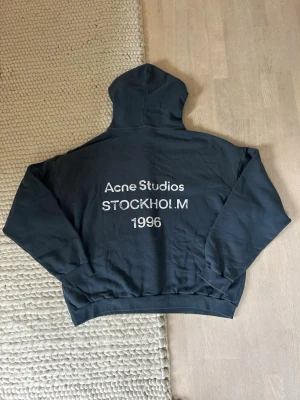  hoodie Acne Studios - Snygg mörkblå hoodie från Acne Studios med tryck på ryggen där det står 'Acne Studios STOCKHOLM 1996'. Klassisk passform med huva och ribbade muddar. Perfekt för en avslappnad och trendig look.