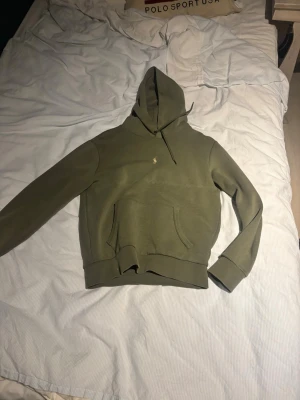 Olivgrön hoodie från Polo Ralph Lauren - Snygg olivgrön hoodie från Polo Ralph Lauren med klassisk broderad logga på bröstet. Hoodien har känguruficka framtill, dragsko i huvan och ribbade muddar vid ärmar och nederkant. Perfekt för en chill och stilren look.