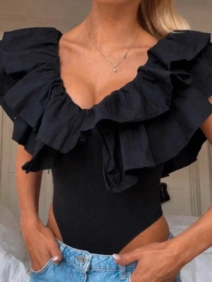 Svart body med volangdetaljer - Trendig svart body med stora volanger längs urringningen och axlarna. Off-shoulder design som ger en snygg och modern look. Tillverkad i stretchigt material som sitter tight mot kroppen. Perfekt att styla med jeans eller kjol.