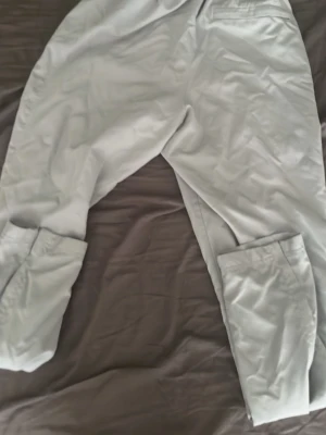 Ljusgrå mjukisbyxor från Under Armour - Snygga ljusgrå mjukisbyxor från Under Armour med elastisk midja och dragsko. Byxorna har meshfoder och raka ben, perfekta för chill eller träning. Sköna och lätta att röra sig i, passar dig som gillar sportig stil.