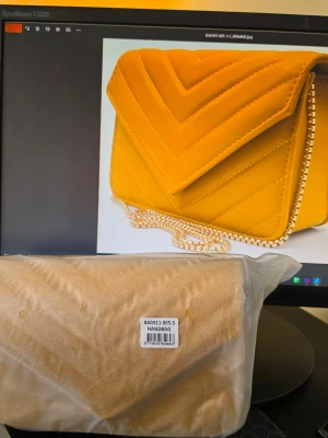 Gul quiltad handväska med kedja - Trendig gul handväska i quiltad design med V-mönster och guldfärgad kedjerem. Väskan har en kuvertformad lockstängning och är tillverkad i ett mjukt material som ser ut som sammet eller mocka. Perfekt för dig som vill sticka ut med färg och stil.