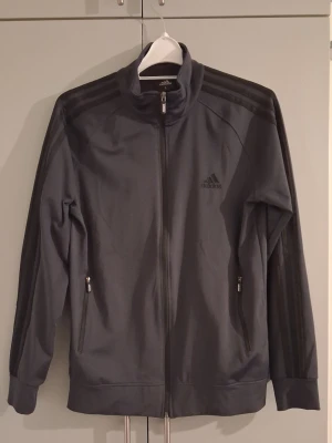 Svart träningsjacka från Adidas - Snygg mörk marinblå träningsjacka från Adidas med hel dragkedja, ståkrage och klassiska tre ränder längs ärmarna. Två fickor med dragkedja och Adidas-logga på bröstet. Perfekt för träning eller som sportig vardagsstil. Det passar storlek M/L