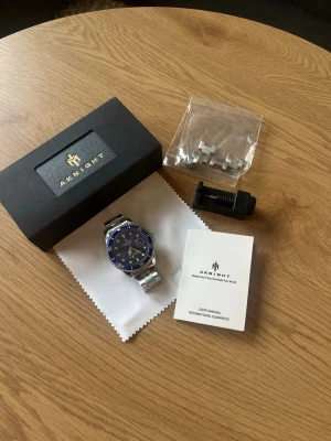 Blå Aknight klocka - Snygg klocka från Aknight med blå urtavla och silverfärgat armband i metall. Klockan har tydliga markeringar, datumfönster och vridbar bezel. Levereras med extra länkar, verktyg, putsduk, originalask och manual.