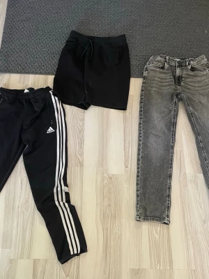 Klädpaket med 3 par byxor, storlek 146–152 - Säljer ett fint klädpaket med 3 par byxor:  • Svarta shorts, storlek 146 • Grå jeans, storlek 152 • Svarta Adidas träningsbyxor med vita ränder, storlek 152  Alla är i fint skick och passar perfekt till vardag, träning och fritid.