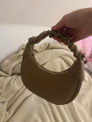 handväska  - Snygg beige handväska i halvmåneform med scrunchie-inspirerat handtag. Väskan är gjord i ett slätt, konstläderliknande material och har dragkedja upptill. Perfekt accessoar för att lyfta din outfit.