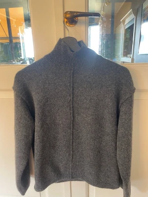 Grå stickad polotröja från H&M - Mysig grå stickad polotröja från H&M med hög krage och rak passform. Tröjan har en markerad söm framtill och långa ärmar. Perfekt för kyliga dagar och enkel att matcha med olika outfits.