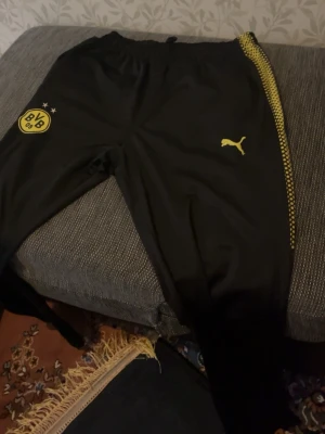 BVB träningsset från Puma - Säljer ett BVB Borussia Dortmund träningsset från Puma. Setet består av svarta träningsbyxor med gula detaljer och klubbmärke samt en gul långärmad träningströja med halv dragkedja och svarta grafiska mönster på sidorna. Perfekt för fotbollsträning eller gym.