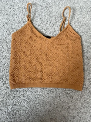 Brun ribbad stickad crop top - Säljer en brun stickad crop top med smala axelband och ribbad kant nedtill. Toppen har ett snyggt geometriskt mönster i materialet och justerbara band för bästa passform. Perfekt att styla med jeans eller kjol.