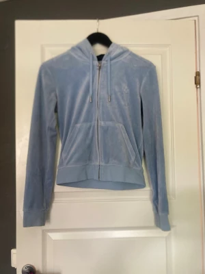 Ljusblå velour hoodie med dragkedja - Säljer en ljusblå hoodie i mjuk velour med dragkedja framtill och två fickor. Tröjan har huva med snören och ett diskret broderat monogram på bröstet. Perfekt för en chill och avslappnad stil.