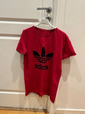 Röd Adidas t-shirt med logga - Snygg röd t-shirt från Adidas med stor svart Trefoil-logga på bröstet. Klassisk rund halsringning och korta ärmar. Tillverkad i mjuk bomull som känns skön mot huden. Perfekt för en sportig och avslappnad stil.
