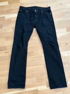 Svarta Levi's 501 jeans straight fit - Klassiska svarta Levi's 501 jeans med rak passform och fem fickor. Jeansen har knappgylf, nitar vid fickorna och den ikoniska läderpatchen bak i midjan. Tillverkade i kraftig denim med snygg svart tvätt. Perfekta för en stilren och tidlös look.