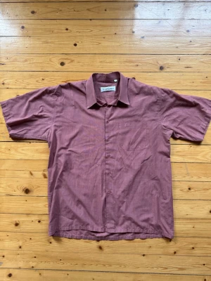 Yves Saint Laurent skjort - Säljer en klassisk rutig skjorta från Yves Saint Laurent i en mörkröd nyans. Skjortan har korta ärmar, knappar framtill och en bröstficka med diskret YSL-broderi. Perfekt för dig som gillar stilrena och tidlösa plagg.