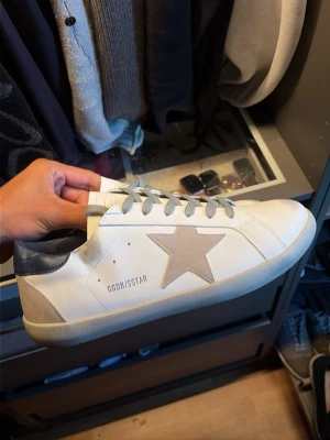 Golden goose skor - Snygga vita sneakers med grå stjärndetalj på sidan och grå häl. Skorna har grå skosnören och en klassisk låg profil. Sulan är vit med svart fiskbensmönster undertill. Märket GGDBISSTAR syns på sidan. Perfekta för en trendig streetstyle-look. Ingen box
