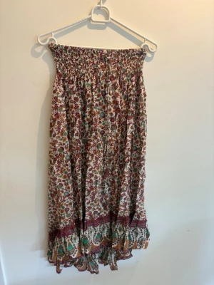 Blommig långkjol med volangkant - Säljer en långkjol med blommigt mönster i vinrött, grönt och beige. Kjolen har smock i midjan och en volangkant längst ner. Perfekt för dig som gillar en avslappnad och färgglad stil.