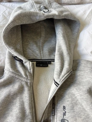 Ljusgrå hoodie från Sail Racing - Ljusgrå hoodie från Sail Racing med dragkedja framtill, broderad logga på bröstet och huva med svart/vita detaljer. Mjuk fleece på insidan och praktisk bröstficka med dragkedja. Perfekt för chill dagar och streetwear-stil.