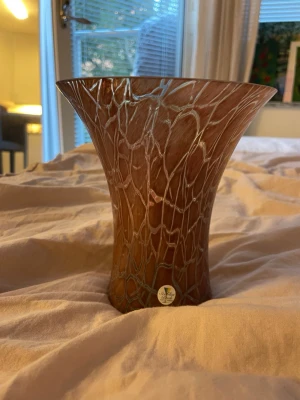 Brun glasvas med mönster - Unik brun glasvas med organiskt, nätliknande mönster i vitt. Vasen har en bred öppning och smalare fot, vilket ger en elegant form. Perfekt som dekoration eller för blommor. Tillverkad i glas och har en dekorativ etikett på foten.
