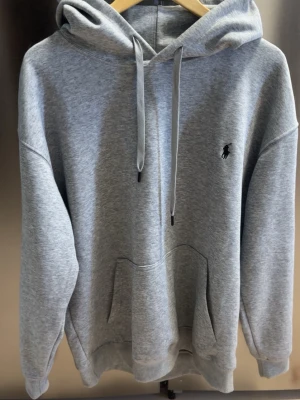 Grå hoodie från Polo Ralph Lauren - Snygg grå hoodie från Polo Ralph Lauren med klassisk huva och dragsnören. Tröjan har en stor magficka och den ikoniska lilla broderade loggan på bröstet. Tillverkad i mjukt sweatshirtmaterial som är perfekt för chill dagar.