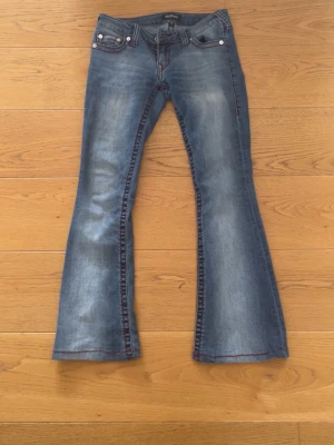 Blå bootcut jeans True Religion - Säljer ett par blå bootcut jeans från True Religion med röda kontrastsömmar och klassisk femficksmodell. Jeansen har en snygg tvätt och utsvängda ben som ger en retrovibe. Perfekta för dig som gillar statement-jeans och vill sticka ut lite extra. Säljer då de tyvärr är för stora