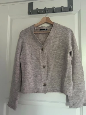 Beige kofta från 157 med knappar - Mysig beige kofta från 157 med tre stora knappar framtill. Koftan har v-ringad hals, lång ärm och är stickad i ett mjukt material. Perfekt att slänga över en t-shirt eller topp för en avslappnad look.