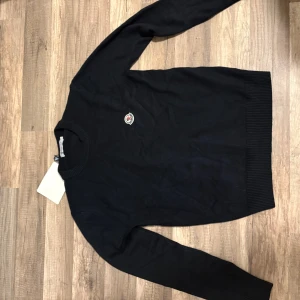 Svart Moncler stickad tröja - Snygg mörkblå stickad tröja från Moncler med rund halsringning och diskret logga på bröstet. Tröjan har ribbade muddar vid ärmslut och nederkant samt långärmad passform. Perfekt för dig som gillar stilrena och exklusiva plagg. 