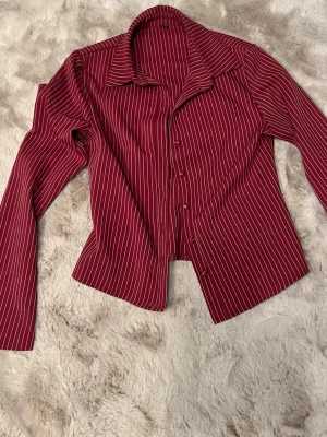 Vinröd randig skjorta med knappar - Säljer en vinröd skjorta med vita ränder och klassisk krage. Skjortan har långa ärmar och knäppning framtill. Perfekt för dig som gillar en stilren look med lite edge.