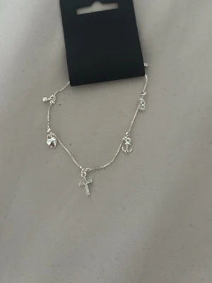 Silverfärgat armband med berlocker pilgrim - Snyggt silverfärgat armband med flera berlocker, bland annat ett kors, ett ankare, ett hjärta och en liten bricka. Helt nytt och oanvänt