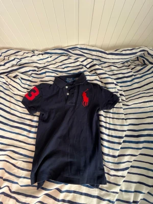 Mörkblå Ralph Lauren pikétröja  - Mörkblå pikétröja från Polo Ralph Lauren med röd logga på bröstet och stor röd siffra 3 på ärmen. Klassisk pikékrage, knappar framtill och slim passform. Tillverkad i mjuk bomull för en clean och sportig vibe.Riktigt fräsch sommar plagg.